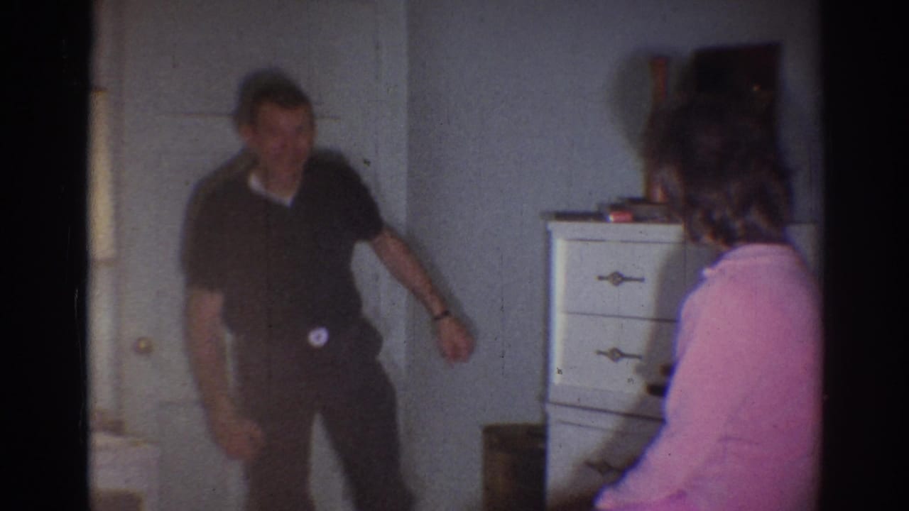 The man appears mid-gesture in a dim 1967 Los Angeles interior, with a woman in pink seated or turned toward him near a dresser.