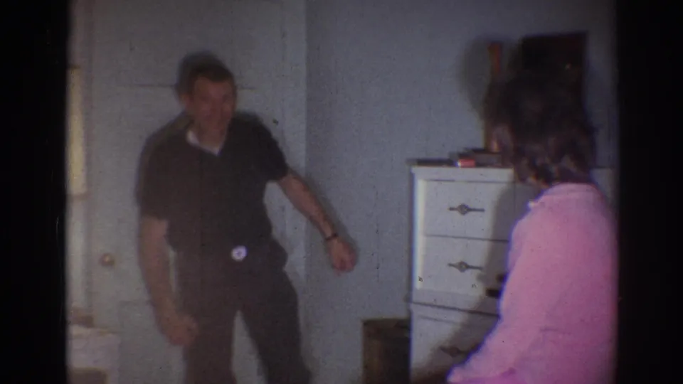 The man appears mid-gesture in a dim 1967 Los Angeles interior, with a woman in pink seated or turned toward him near a dresser.