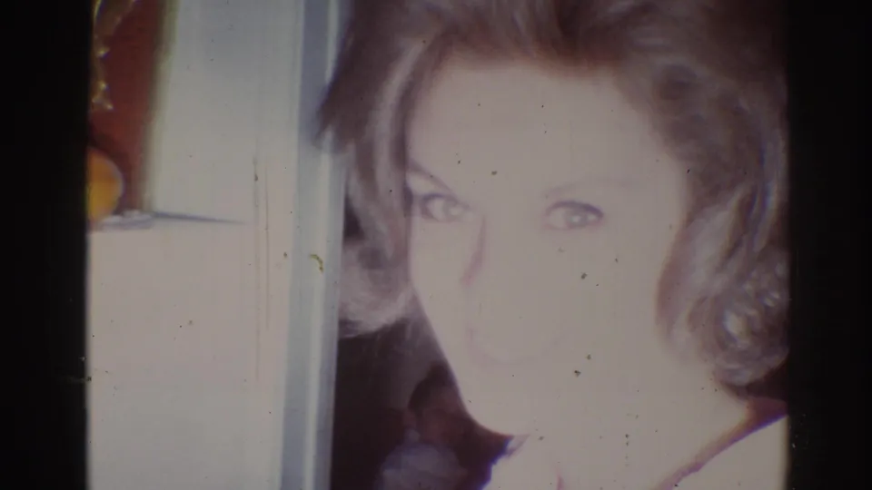 1967 Los Angeles home-movie frame of a blonde woman smiling toward the camera in a private indoor setting.