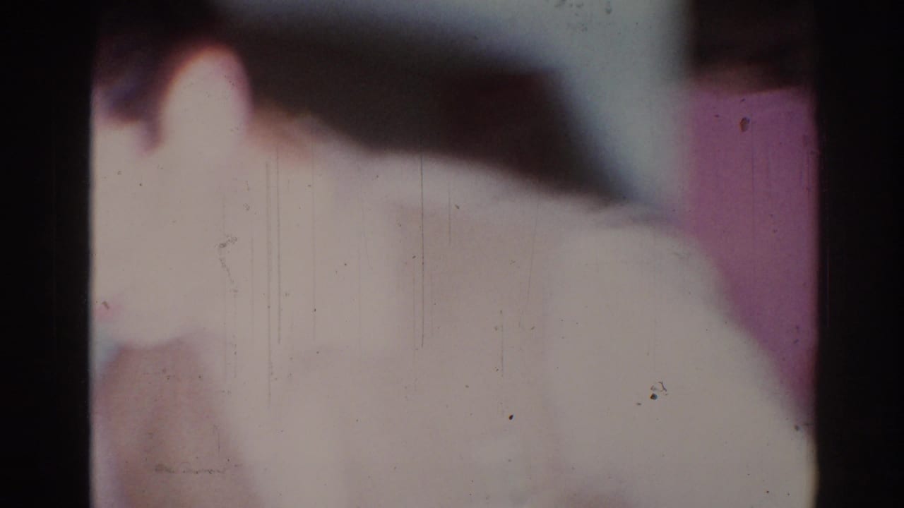 Soft, overexposed 8mm frame showing a person passing close to the camera inside a private room.