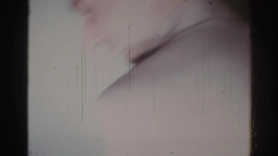 Home-movie frame with a blurred adult figure in a light shirt moving close to the lens inside a private interior.