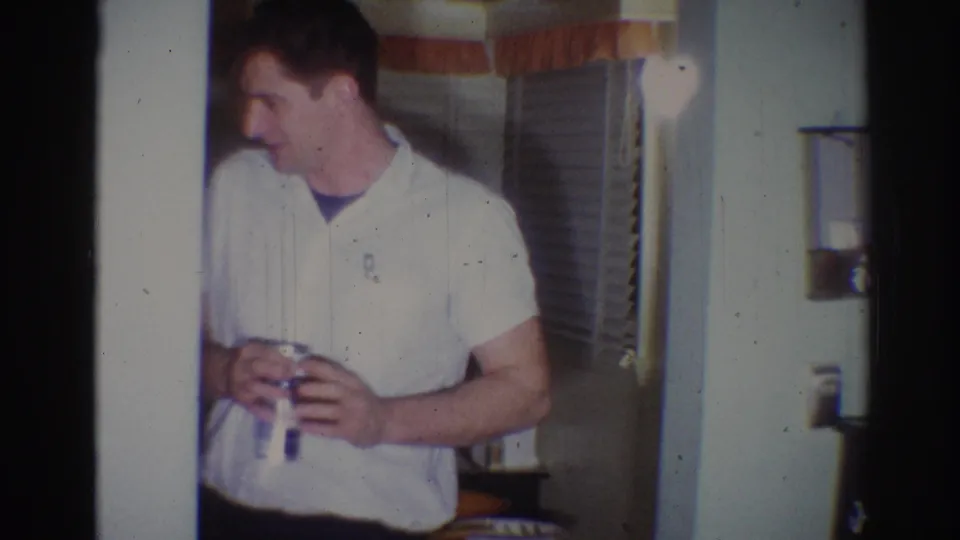 1967 Los Angeles home-movie frame of a man in a white short-sleeved shirt holding a drink near curtains and blinds.