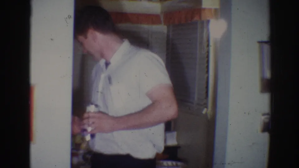 In a 1967 Los Angeles home-movie frame, a young man in a white shirt holds a beer can beside a doorway and drawn blinds.