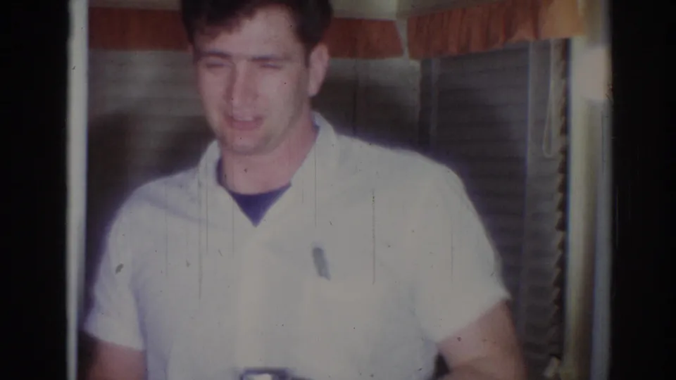 Closer 8mm frame of the young man in a white short-sleeved shirt, with drawn blinds and warm interior light behind him.