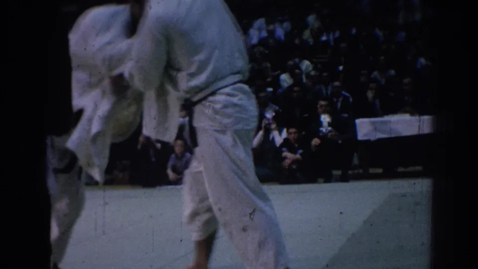 Two martial arts competitors in white gi struggle on an indoor mat before spectators in Los Angeles footage from 1967.