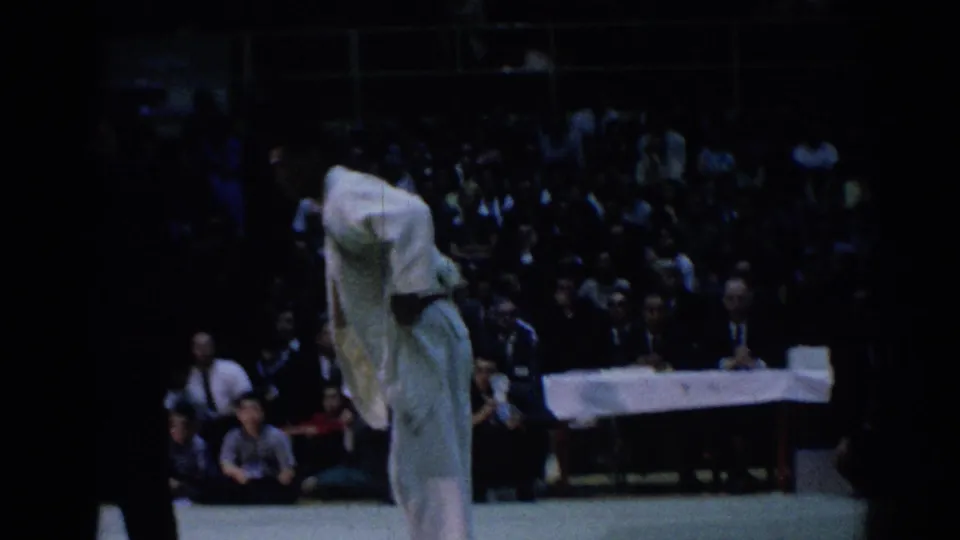 A competitor in a white gi bends low during a judo match as spectators and an officials table appear in the background.