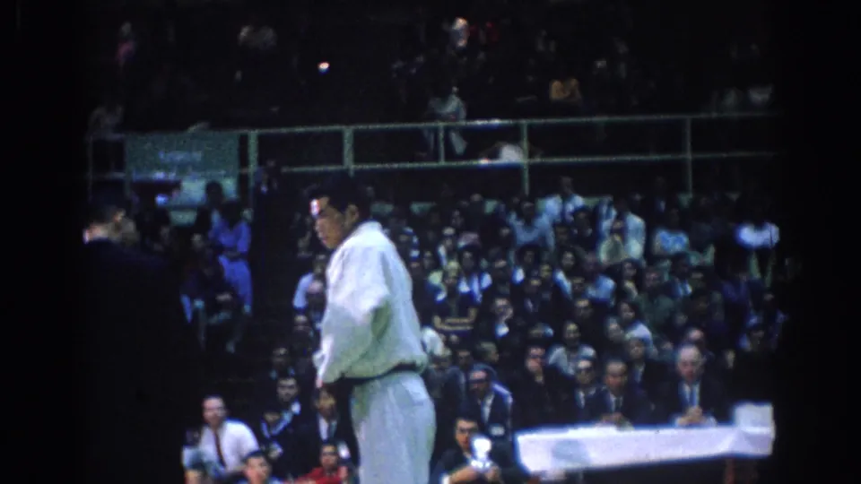 A martial arts competitor in a white gi stands on an indoor mat before a crowded spectator area in 1967 Los Angeles footage.