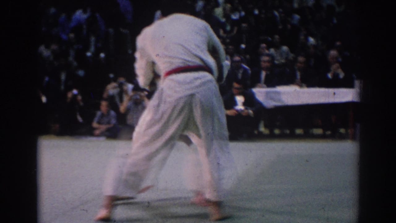 1967 Los Angeles frame of a karate competitor in a white gi with a red belt on a light mat, with judges and spectators behind.