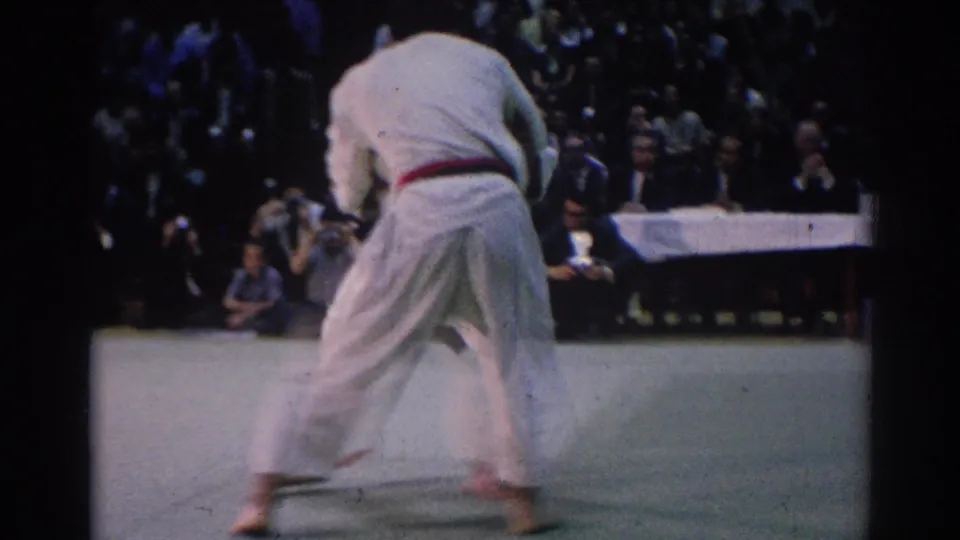 1967 Los Angeles frame of a karate competitor in a white gi with a red belt on a light mat, with judges and spectators behind.