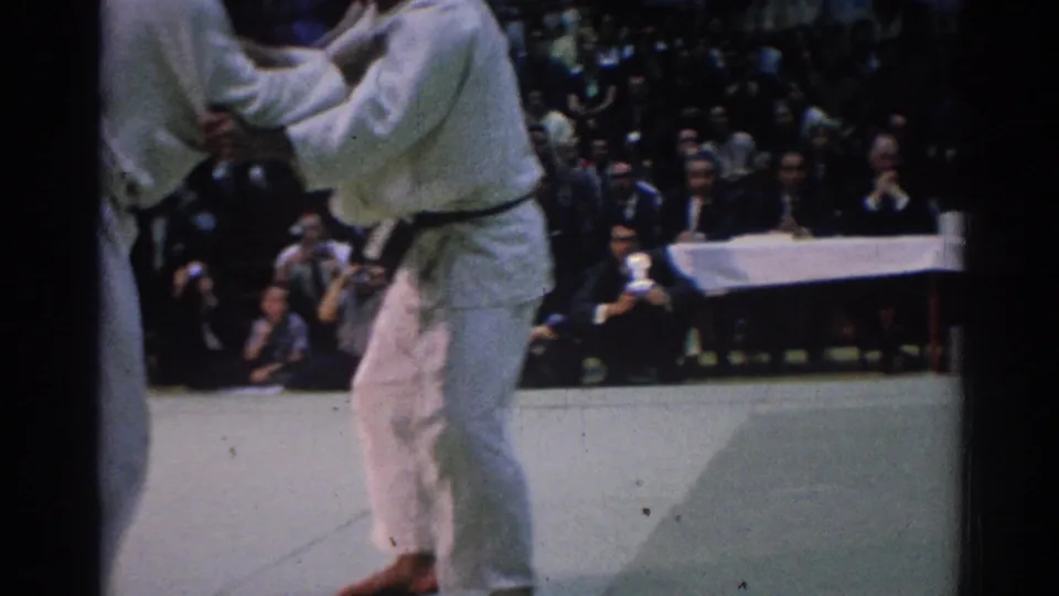 White-gi competitors continue an upright grapple in the Los Angeles contest, with photographers and spectators visible behind them.