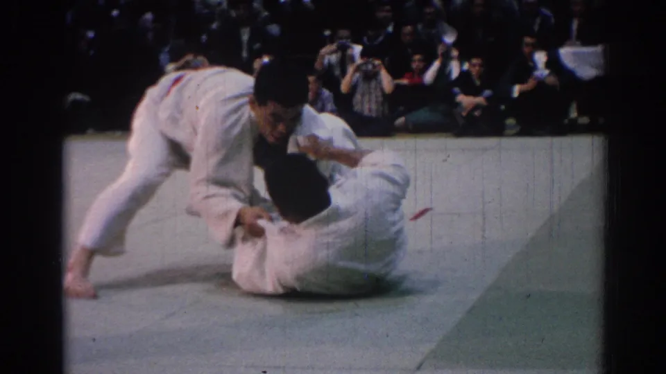 A 1967 Los Angeles judo-style exchange shows one competitor on the mat and the other leaning over him as spectators look on.