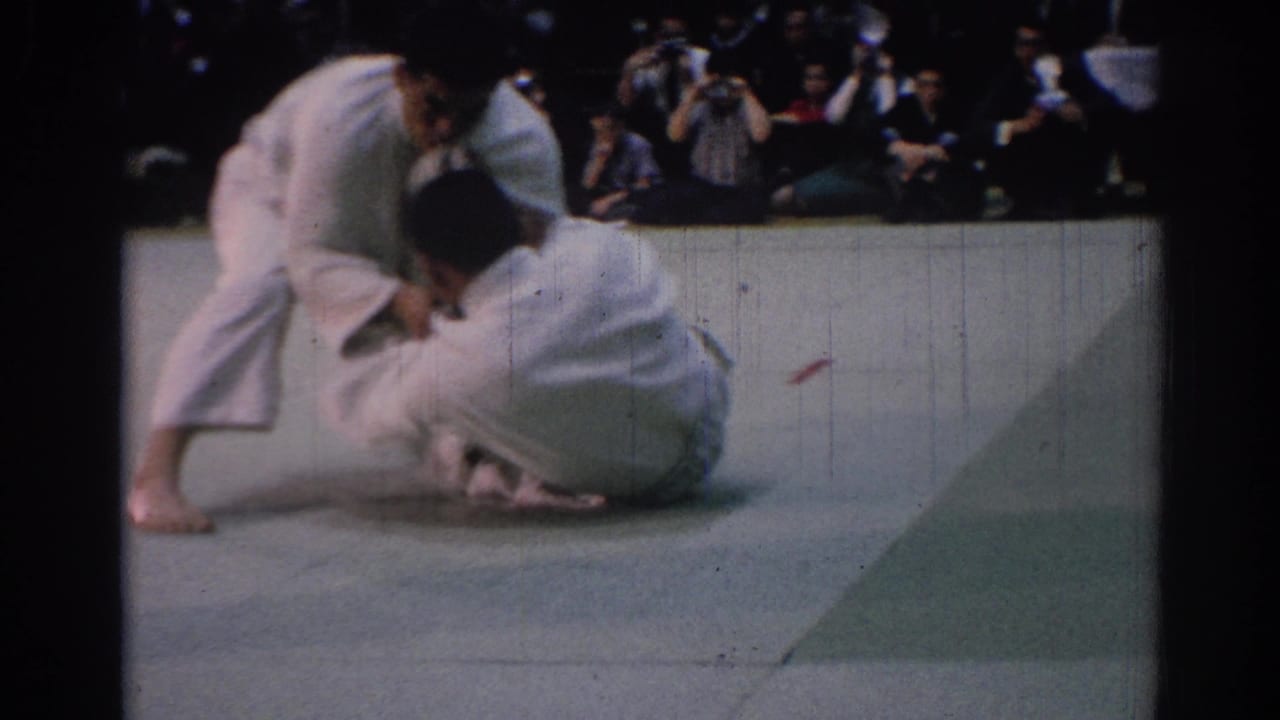 Two men in white gi grapple low on a mat while spectators sit behind them.