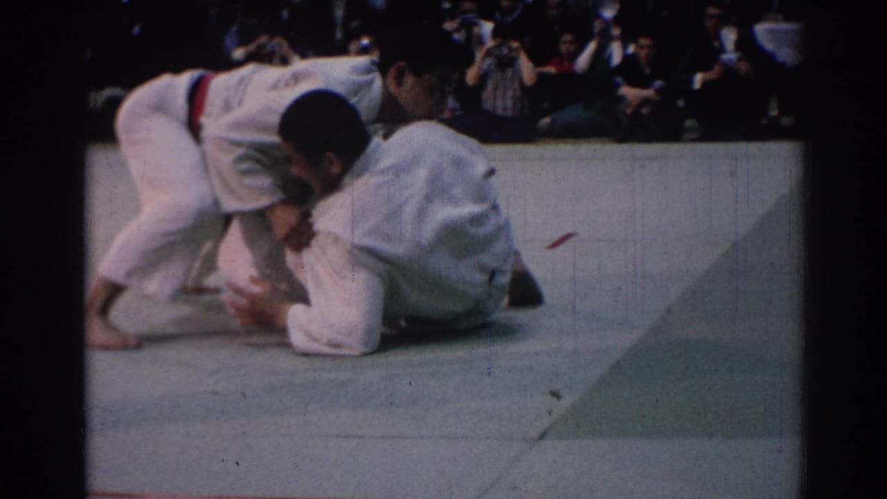 1967 Los Angeles 8mm frame showing one judo participant kneeling or bracing while another in a white gi leans over him during the match.