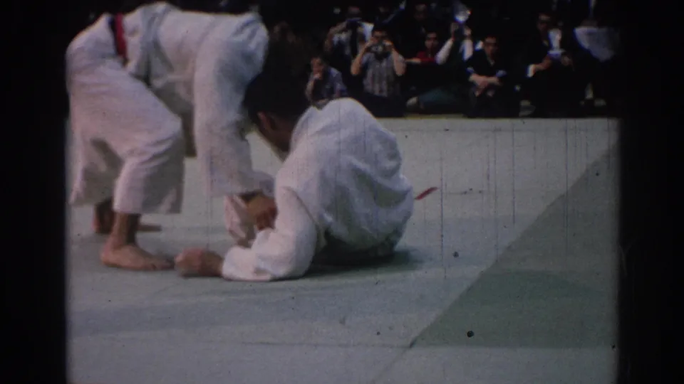 1967 Los Angeles 8mm frame of a judo participant seated on the mat while another white-clad competitor moves at the edge of the frame.