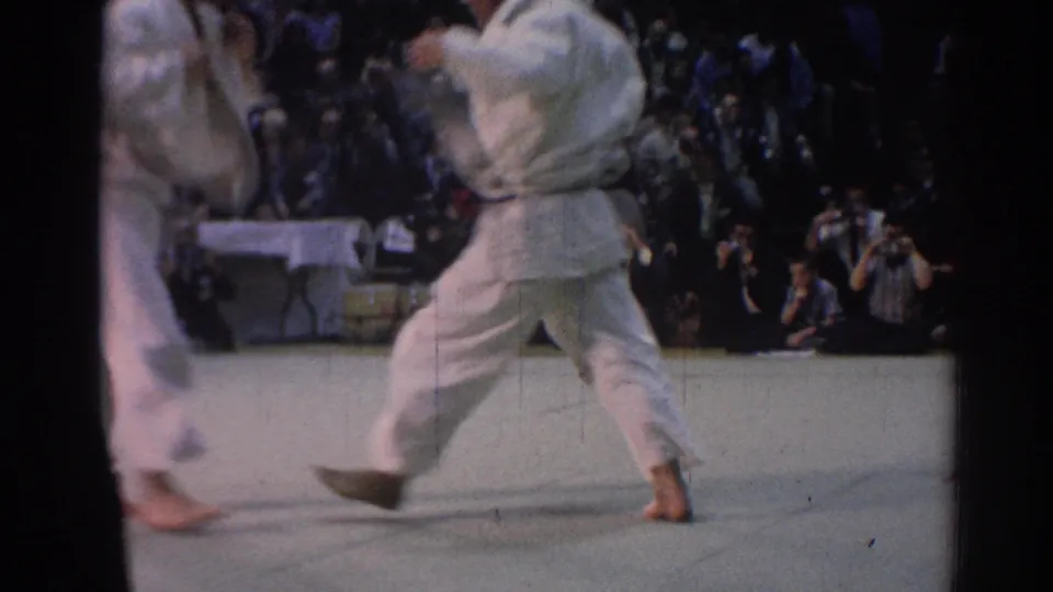 1967 Los Angeles 8mm frame showing a white-gi judo participant stepping across the mat, with a seated audience and photographers in the background.