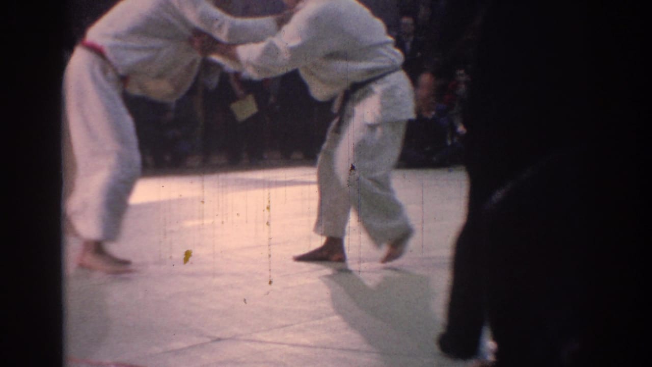 Two karate practitioners in white gi grapple on a mat before spectators.