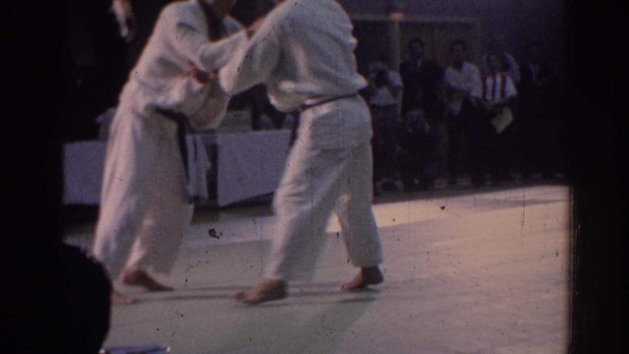 1967 Los Angeles frame with two black belt practitioners in white gi clinching during a karate match in an indoor venue.