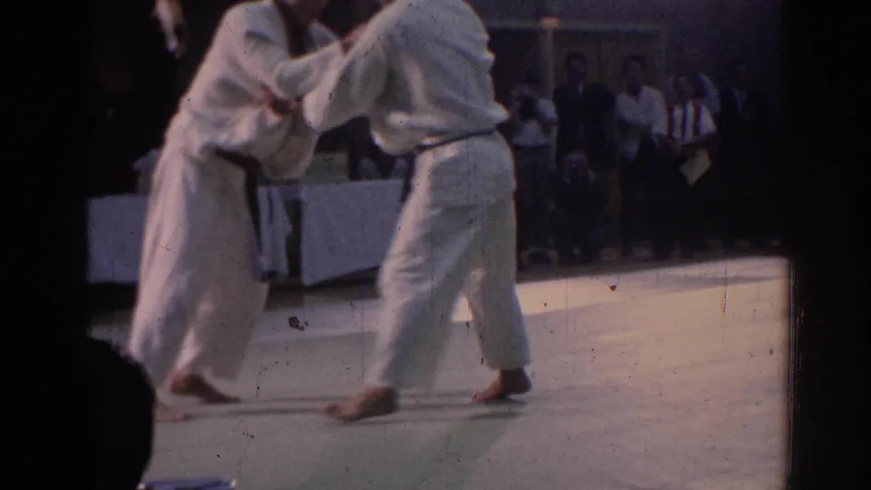 1967 Los Angeles frame with two black belt practitioners in white gi clinching during a karate match in an indoor venue.