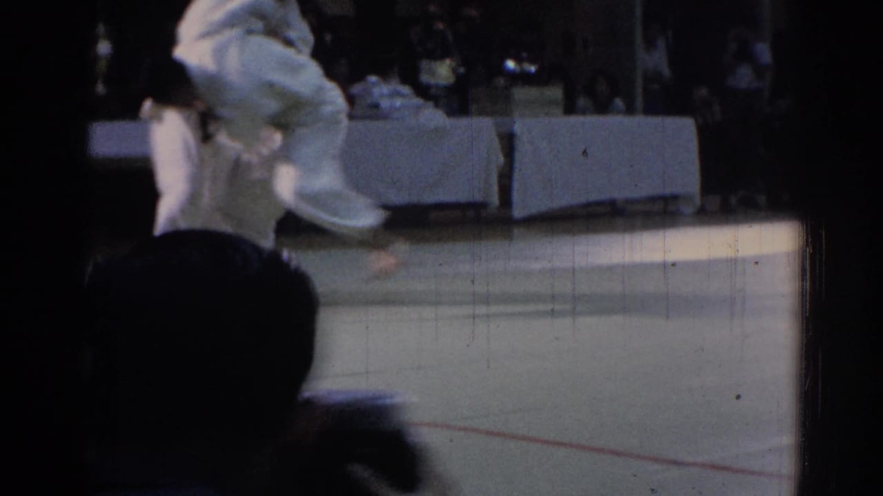 Judo competitors in white gi move on a mat before judges and spectators.