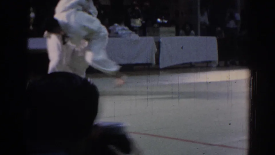 1967 Los Angeles home-movie frame showing judo competitors in white gi on a competition mat with spectators in the background.