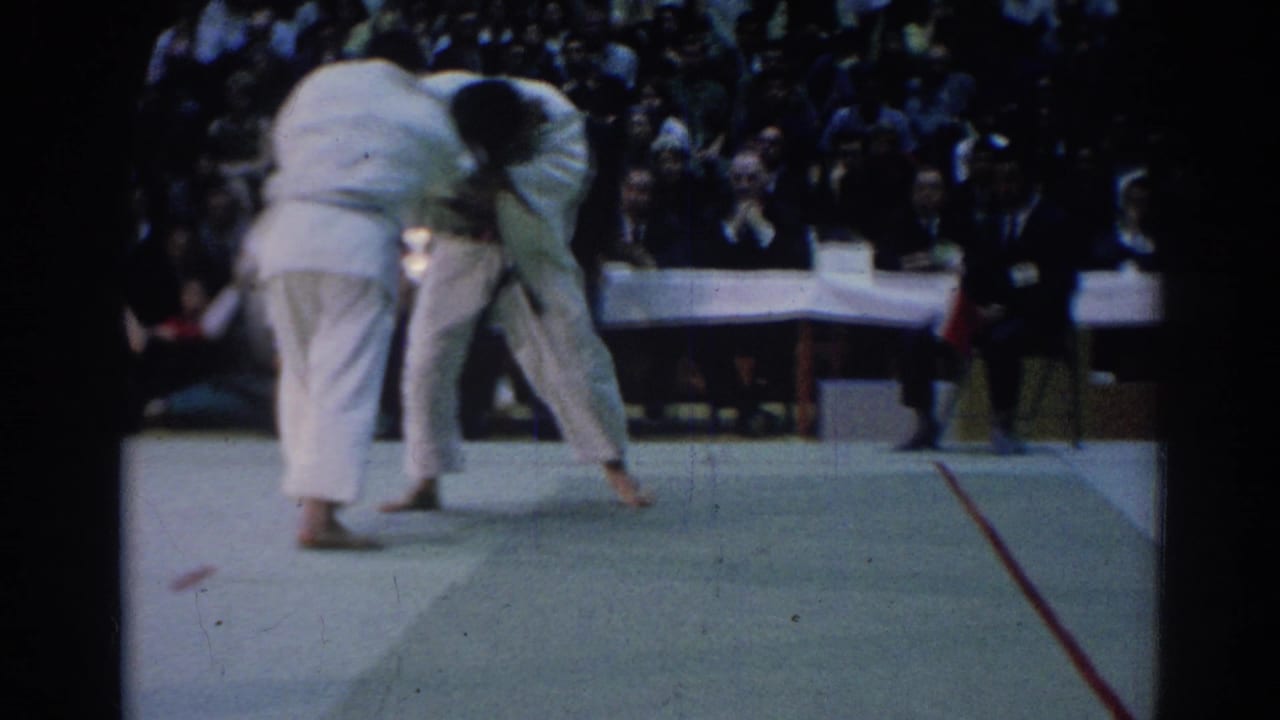 Two judo competitors grapple near the center of a mat with a crowd behind them.