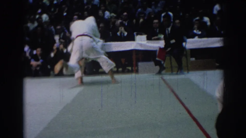 1967 Los Angeles judo match frame showing two black-belt competitors grappling while judges with red flags observe nearby.