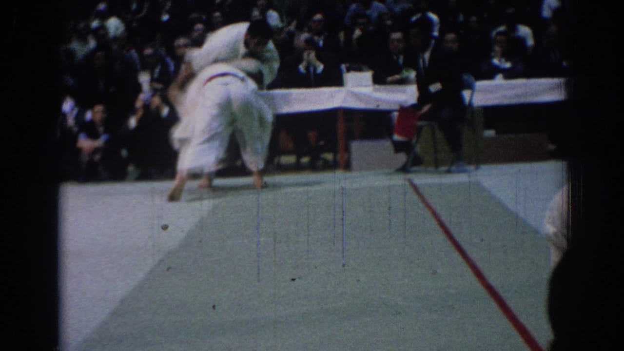 Two judo athletes in white gi uniforms move on a mat as spectators watch behind them.