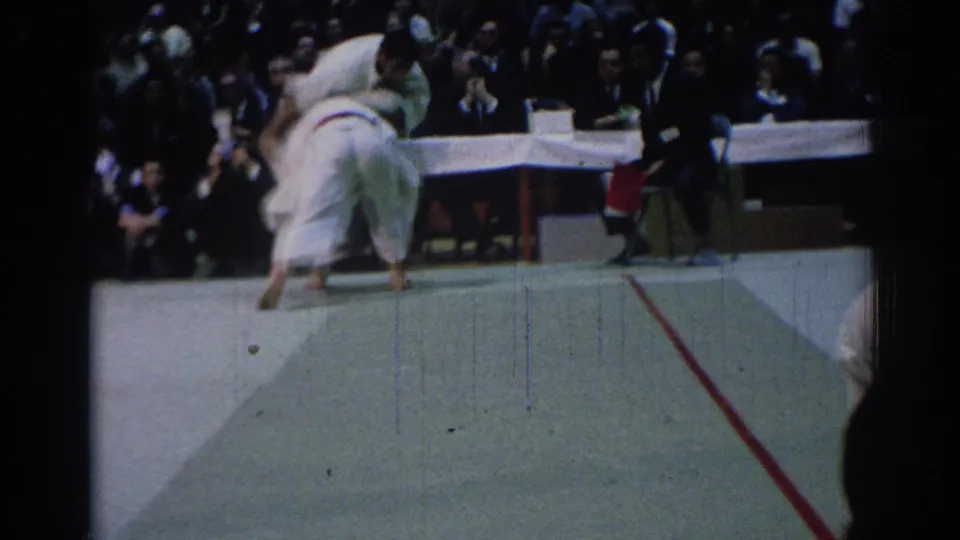 1967 Los Angeles 8mm frame of two judo athletes in white gi uniforms engaged near the edge of a competition mat with spectators behind.