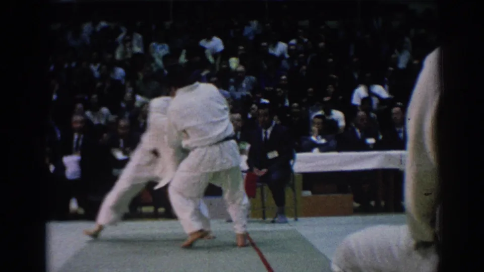 A handheld 8mm frame shows two judo athletes locked in close contact before spectators and officials during the Los Angeles match.