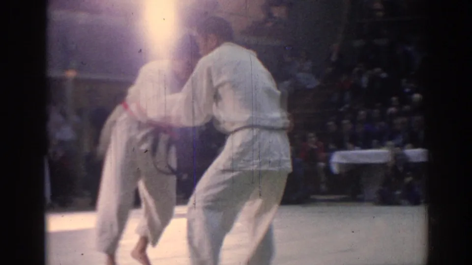 The closing 1967 Los Angeles frame shows two judo athletes in white gi uniforms standing close on the mat with a bright exposure flare above them.