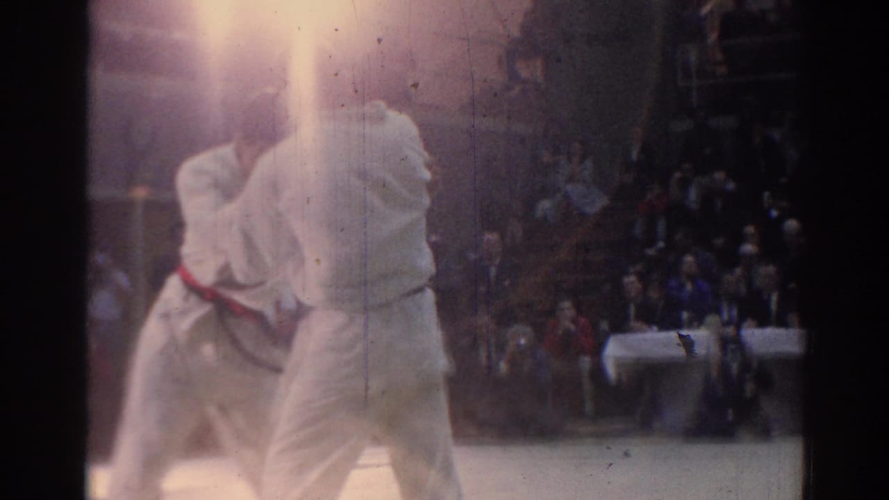 In 1967 Los Angeles footage, two martial artists in white gi and colored belts grapple under bright arena lights.