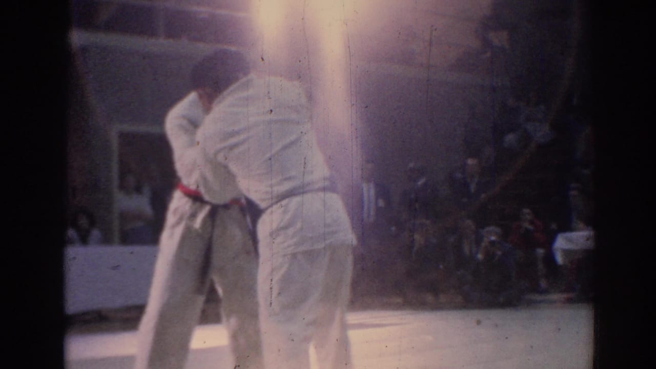 Two martial artists in white uniforms continue a close clinch on a light mat, with spectators visible behind them.