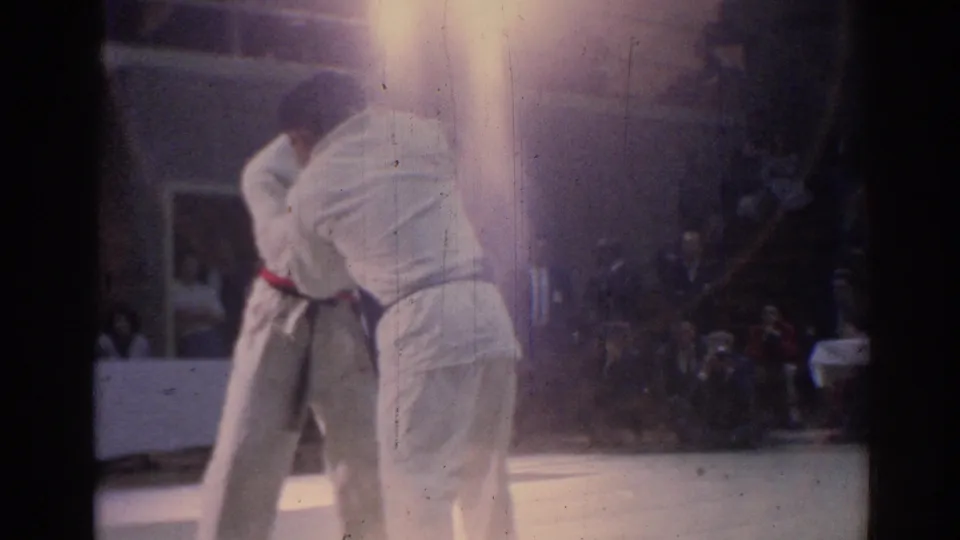Two martial artists in white uniforms continue a close clinch on a light mat, with spectators visible behind them.