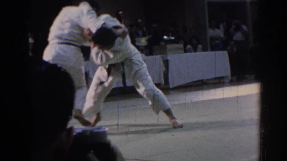 A martial artist drives through a throw or control position while the opponent bends forward on the indoor mat.