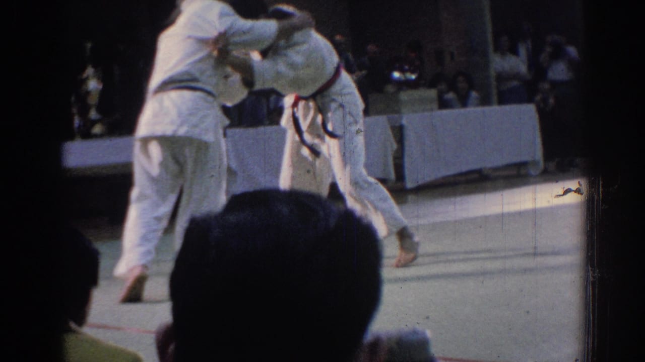 Two martial artists in white gi uniforms grapple on a mat before seated spectators.