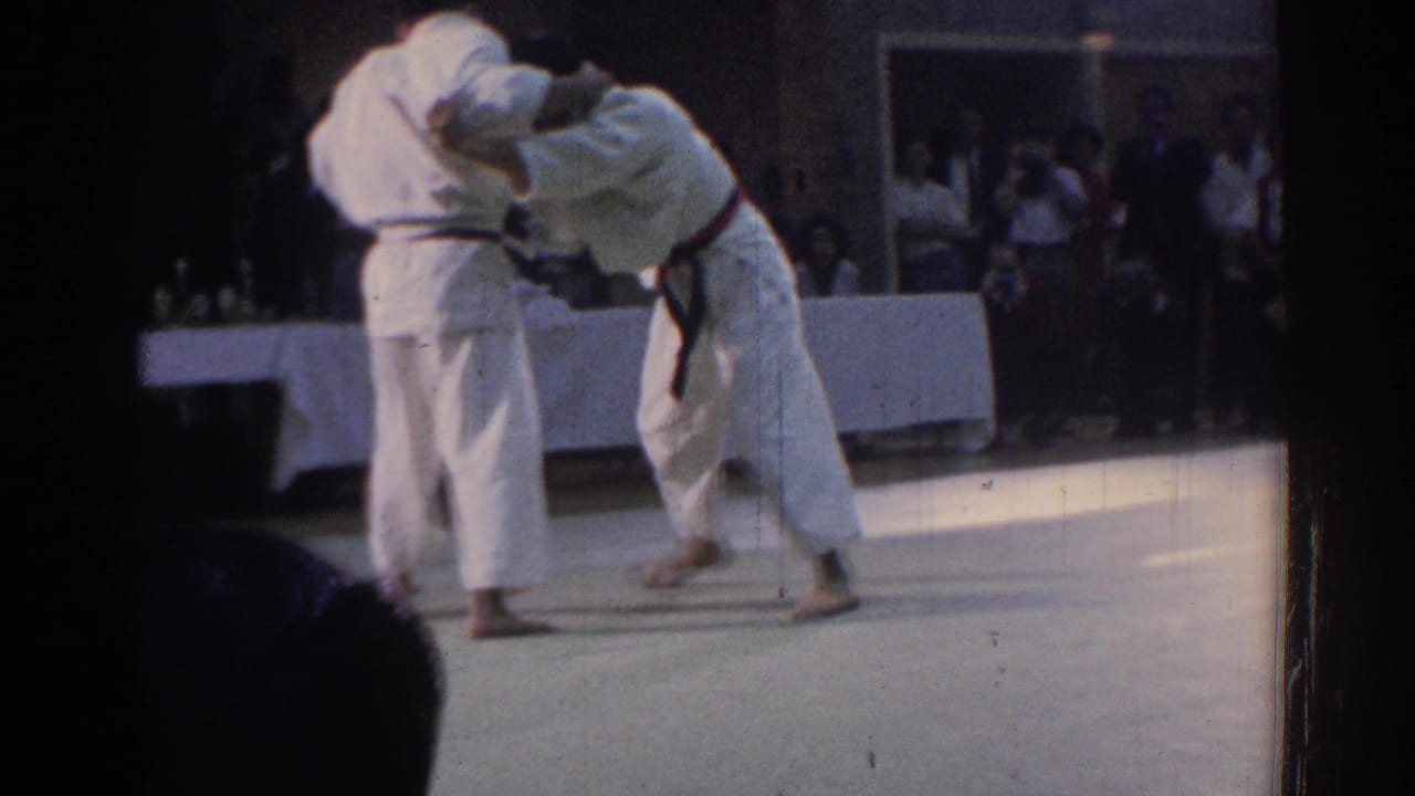 Two martial artists in white uniforms continue a standing grapple on an indoor mat in the 1967 Los Angeles home-movie clip.