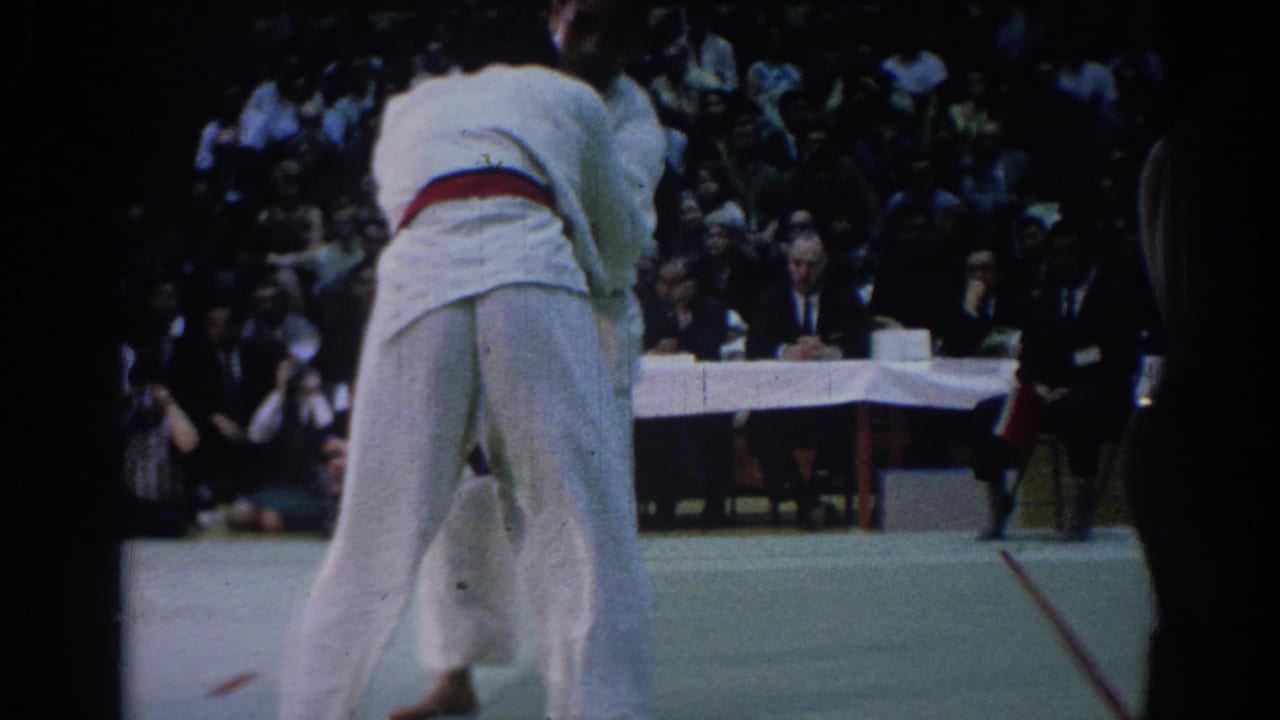 Two gi-clad competitors grapple on a mat in front of judges and spectators.