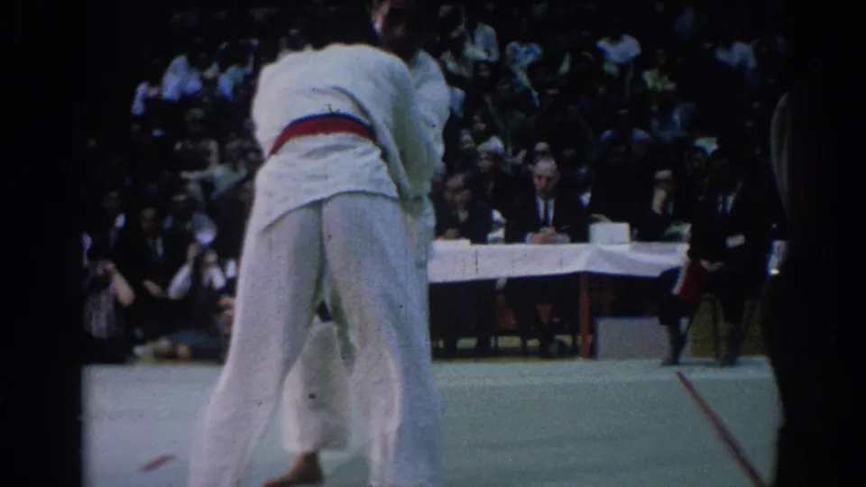 1967 Los Angeles 8mm frame showing two male jiu-jitsu competitors in white gi grappling near judges and a seated crowd.