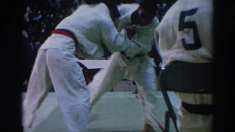 1967 Los Angeles jiu-jitsu frame showing two competitors grappling as an official with a large number on his back stands nearby.