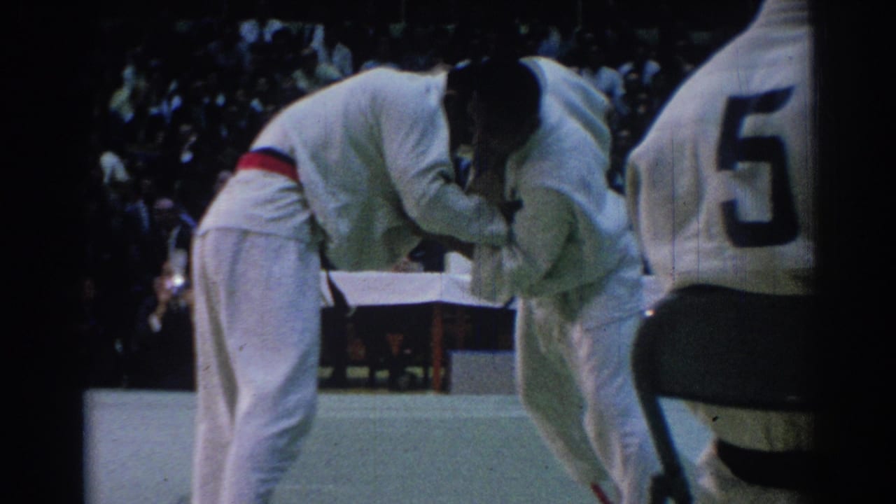 Two martial arts competitors in white gi lean together on an indoor mat as spectators sit behind them.