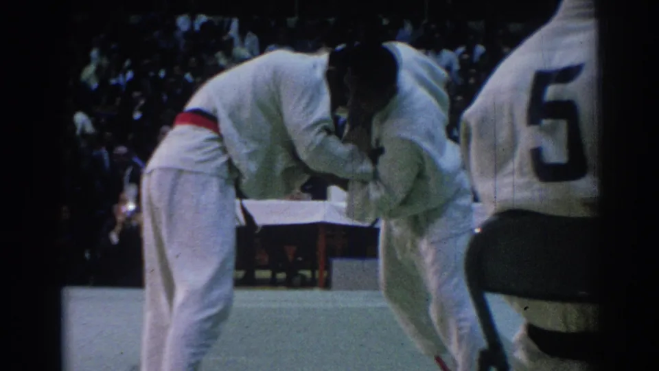 1967 Los Angeles indoor martial arts bout with two competitors in white gi on a green mat before judges and spectators.