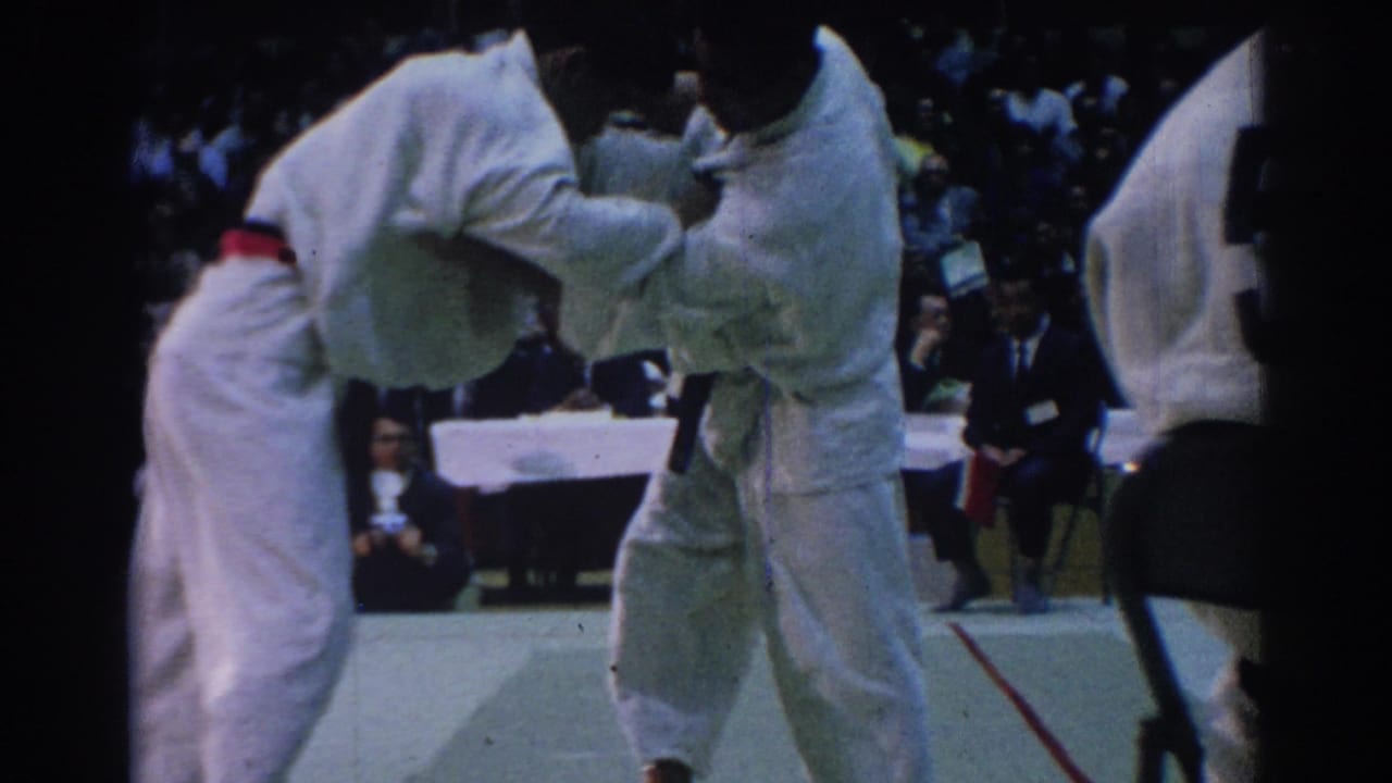 Two white-gi competitors grapple during a 1967 Los Angeles indoor martial arts match, seen from a spectator angle.