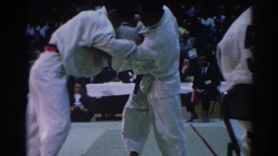 Two white-gi competitors grapple during a 1967 Los Angeles indoor martial arts match, seen from a spectator angle.