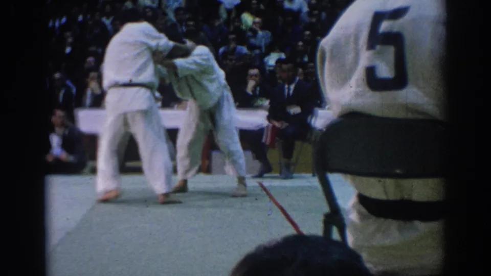 A 1967 Los Angeles martial arts match continues as one competitor bends forward on the mat before seated spectators.