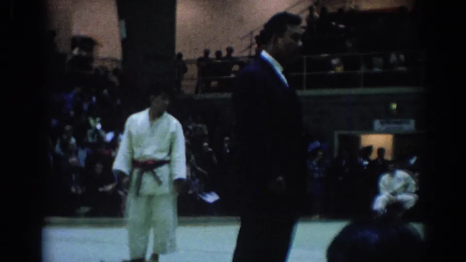 1967 Los Angeles judo footage shows a suited official near a competitor, with spectators seated around the indoor venue.