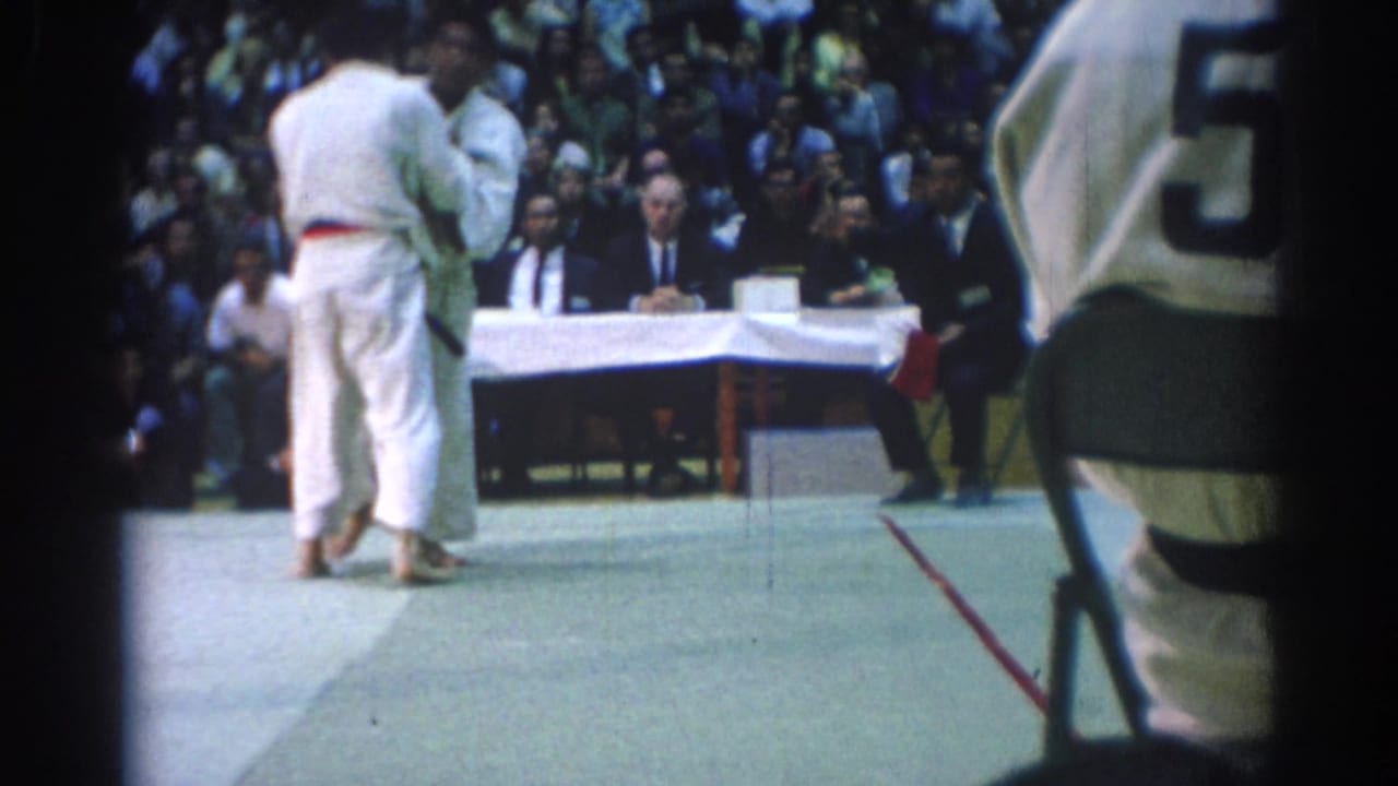 Two judo competitors in white gi stand on a mat before judges and spectators.