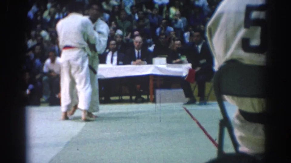 1967 Los Angeles judo competitors in white gi stand near a judges table as a crowd watches from arena seating.