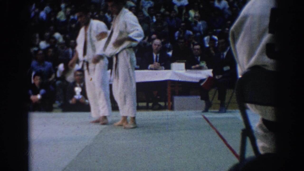 Two men in gi stand on the mat with a judges table and crowd behind them.