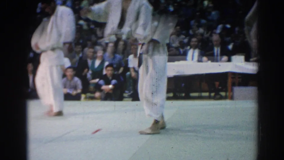 A judo competitor crosses the mat as another athlete waits nearby, with judges and spectators in the background.
