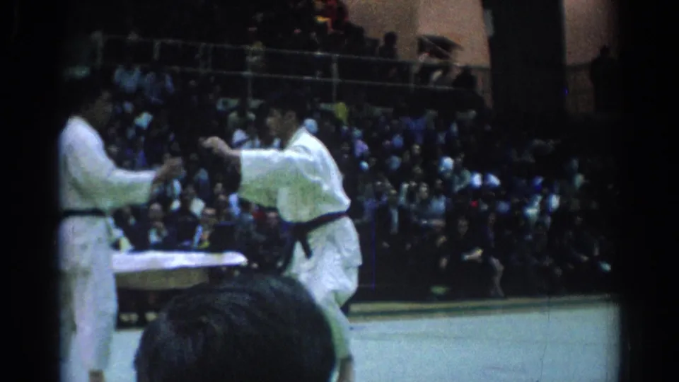 Two judo athletes face each other in a dim indoor arena, with a crowded spectator section and officials nearby.